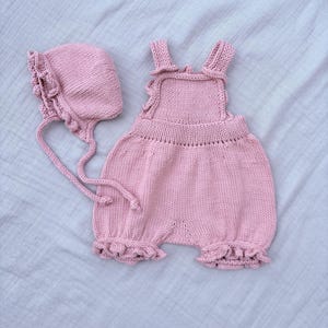 May include: A knitted baby romper and bonnet set in a soft pink color. The romper features ruffled edges and straps, while the bonnet has a matching ruffle trim and ties. The set is made of a soft, textured yarn.
