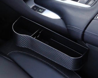 May include: Black car interior organizer with a carbon fiber pattern. The organizer is located in the space between the driver's seat and the center console.