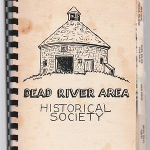 May include: A spiral-bound cookbook with a beige cover featuring a black and white illustration of a round building on a hill. The text "DEAD RIVER AREA HISTORICAL SOCIETY" is printed below the illustration. Tabs on the side indicate recipe categories.