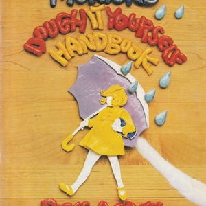 May include: The book cover for Morton's Dough It Yourself Handbook Rises Again features a clay-like relief of a figure in a yellow raincoat holding a purple umbrella under blue raindrops. The title text is rendered in raised, colorful lettering.