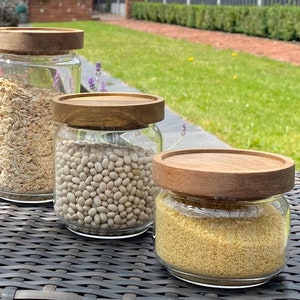 Puede incluir: Tres tarros de vidrio con tapas de madera llenos de diferentes granos. Los tarros están sobre una superficie de mimbre tejido. Los tarros están llenos de avena, frijoles blancos y cuscús.