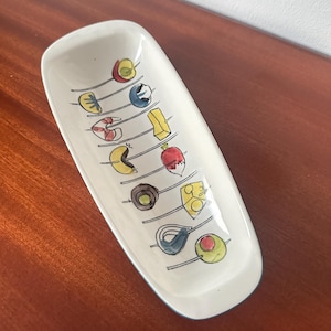 May include: White ceramic serving dish with a blue rim. The dish is decorated with a pattern of colourful food items on skewers, including olives, cheese, and a red radish.