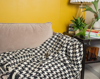Puede incluir: Una manta de tiro de pata de gallo en blanco y negro con flecos en un sofá gris claro en una habitación con una pared amarilla y una lámpara con una pantalla blanca.