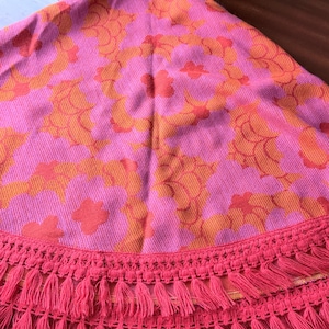 Può includere: Un tessuto circolare caratterizzato da un vivace motivo floreale rosa e arancione. Il tessuto è rifinito con un'ampia bordura decorativa di frange rosa abbinate che pende in più strati lungo il bordo curvo dell'oggetto.