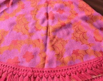 May include: A circular textile featuring a vibrant pink and orange floral pattern. The fabric is finished with a wide, decorative border of matching pink fringe trim that hangs in multiple layers along the curved edge of the piece.