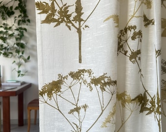 May include: Off-white linen curtains with a repeating pattern of olive green botanical illustrations. The design features detailed depictions of stems, leaves, and flower clusters. The curtains are hanging in front of a window, with a glimpse of a table and greenery visible.