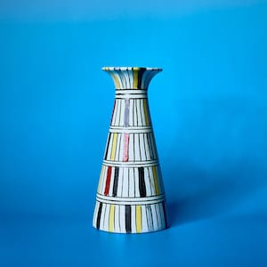 Op de afbeelding: Een decoratieve vaas met een uitlopende bovenkant en een brede basis. De vaas is wit met verticale zwarte strepen en kleurrijke accenten van rood, geel en grijs. De vaas staat tegen een helderblauwe achtergrond.