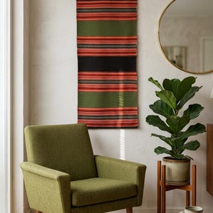 Puede incluir: Un sillón tapizado en color verde musgo con patas de madera, situado bajo un tapiz de pared rectangular y alargado con rayas horizontales en verde oliva, negro y naranja. Un espejo redondo con marco dorado y una planta están cerca.