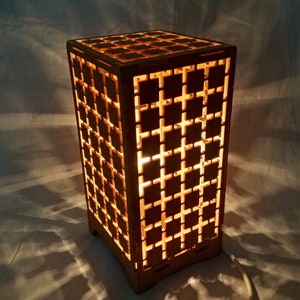 Könnte beinhalten: Eine Holzlampe mit geometrischem Muster, von innen beleuchtet und mit einem warmen Schein. Die Lampe ist aus hellbraunem Holz gefertigt und hat einen quadratischen Sockel.