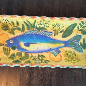 May include: A rectangular ceramic serving dish with a scalloped edge. The dish is painted with a yellow background and features a blue and yellow fish with green foliage and yellow citrus fruit.