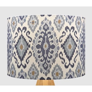 May include: A lampshade with a cream-coloured background and a repeating pattern of blue, grey, and gold geometric shapes. The lampshade is supported by a light-coloured wooden base, creating a decorative lighting fixture.