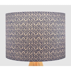May include: A cylindrical lampshade with a repeating geometric pattern in navy blue and cream. The lampshade sits on a light-coloured wooden base, creating a contrast in textures and colours. The design is modern and suitable for home decor.
