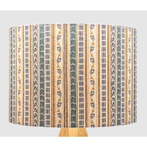 May include: A lampshade with a repeating vertical pattern in beige, teal, and brown. The design features floral and geometric elements. The lampshade is mounted on a light-coloured wooden base. The lampshade is cylindrical.