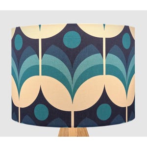May include: A lampshade with a repeating floral design in teal, cream, and various shades of blue. The pattern features stylized flower shapes and circular accents against a dark blue backdrop. The lampshade is supported by a light-coloured wooden base.