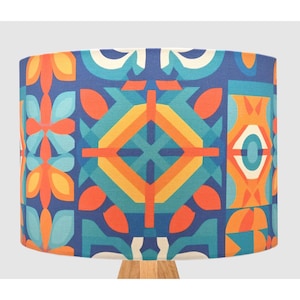 May include: A cylindrical lampshade with a geometric pattern in shades of blue, orange, yellow, and white. The design features stylized floral and geometric shapes. The lampshade sits on a wooden base, creating a decorative lighting fixture.