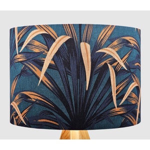 May include: A cylindrical lampshade featuring a dark teal background with a repeating pattern of large, elongated palm leaves in shades of navy blue and light tan. The fabric has a visible woven texture throughout the entire surface area.