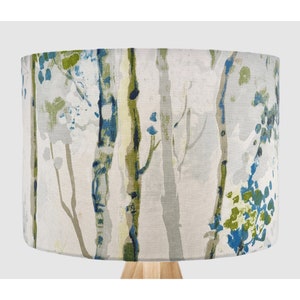 May include: A cylindrical lamp shade featuring a printed pattern of vertical tree trunks in shades of blue, green, and grey against a light off-white background. The fabric texture is visible across the surface of the drum-shaped lamp shade.
