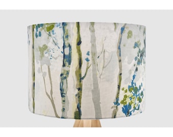 May include: A cylindrical lamp shade featuring a printed pattern of vertical tree trunks in shades of blue, green, and grey against a light off-white background. The fabric texture is visible across the surface of the drum-shaped lamp shade.