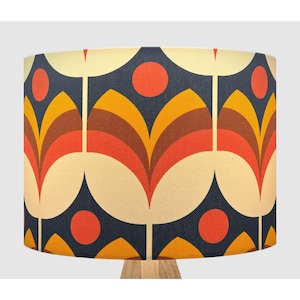 May include: A cylindrical lampshade featuring a repeating pattern of stylised tulip-like shapes in shades of orange, yellow, and brown against a dark navy blue background, accented with solid orange circles positioned between the floral motifs.
