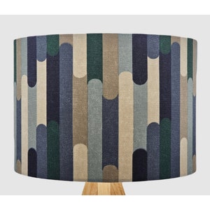 May include: A cylindrical lampshade featuring a repeating pattern of vertical, rounded-end stripes in shades of navy blue, forest green, light grey, and beige. The fabric has a visible woven texture, and the top of a wooden lamp base is visible below.