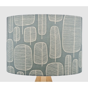 May include: A cylindrical lampshade featuring a blue-grey fabric background with a repeating pattern of white, hand-drawn style tree silhouettes. The trees are depicted with simple vertical trunks and rounded, leaf-filled canopy outlines.