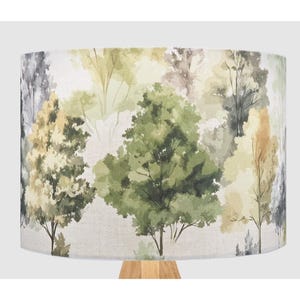 May include: A cylindrical lampshade with a watercolour forest design. The artwork depicts trees in shades of green, yellow, and grey against a neutral backdrop. The lampshade is mounted on a light-coloured wooden base.