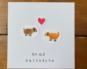 Puede incluir: Una tarjeta blanca con una ilustración de un dachshund marrón y naranja, un corazón rosa y el texto "be my valentine".