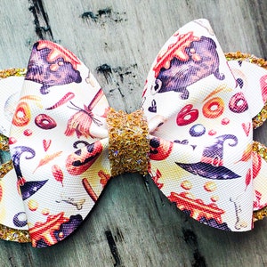 Puede incluir: Un lazo blanco con un estampado de temática de Halloween que presenta brujas, calderos, calabazas y maíz dulce. El lazo está adornado con purpurina dorada.