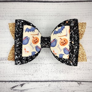 May include: Black and gold glitter hair bow with a Halloween-themed print featuring cauldrons, bats, skulls, and pumpkins.