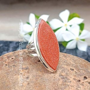 Pode incluir: Um anel de prata com uma pedra grande, alongada, castanho-avermelhada. A pedra tem uma aparência texturizada e brilhante e é colocada em uma moldura de prata. O anel é exibido em uma rocha com flores brancas ao fundo. Etsy @ Unique Silver Crafts.