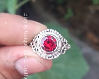 Puede incluir: Un anillo de plata con una piedra preciosa roja engastada en un diseño de filigrana decorativo. El anillo está en un dedo.
