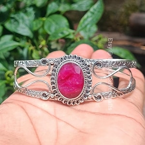 Op de afbeelding: Een zilveren armband met een sierlijk ontwerp met een grote, ovale roze edelsteen. De armband is open en verstelbaar.
