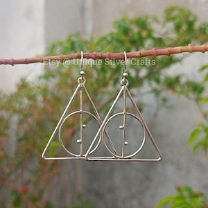 May include: Silver-colored drop earrings featuring the Deathly Hallows symbol. Each earring has a triangle with a circle and a vertical line through it. The earrings are hanging from a branch with green foliage in the background. The text "Etsy @ Unique Silver Crafts" is visible.