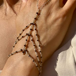 May include: A silver hand chain with small, dark beads. The chain connects at the wrist and extends over the back of the hand, with multiple strands of beads and chain. The jewelry is displayed against a neutral background.