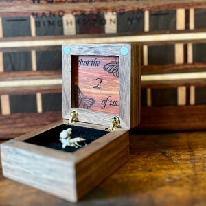 May include: A wooden ring box with a hinged lid. The lid is open and reveals a red velvet interior with the words "Just the 2 of us." engraved on it. Two butterfly designs are also engraved on the lid. The box is made of dark wood and has a light wood inlay. The box is sitting on a wooden surface.