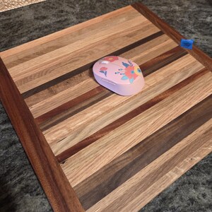 May include: A square wooden cutting board featuring alternating light and dark wood strips arranged in a linear pattern. A light pink computer mouse with a colorful floral print sits on the surface of the board, which has a small blue piece of tape on the edge.