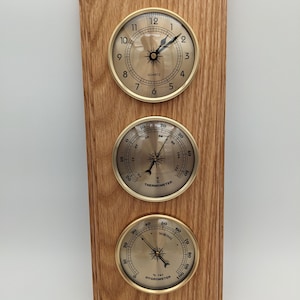 May include: A wooden wall clock with three round dials. The top dial is a clock with black hands and gold numbers. The middle dial is a thermometer with a black needle and a gold face. The bottom dial is a hygrometer with a black needle and a gold face. The dials are all set against a light brown wooden background.