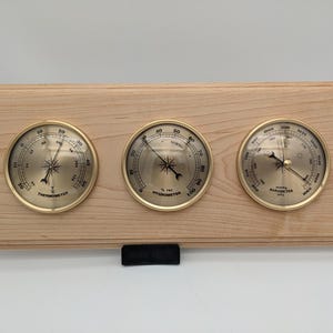 May include: A wooden wall-mounted weather station with three round analog gauges. The gauges are labeled "Thermometer", "Hygrometer", and "Barometer".