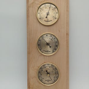 May include: A wooden wall-mounted weather station with three analog gauges. The gauges are for temperature, humidity, and barometric pressure. The gauges have gold-colored bezels and white faces with black markings.