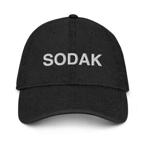 Peut inclure: Une casquette de baseball noire avec le mot "SODAK" brodé en blanc sur le devant.