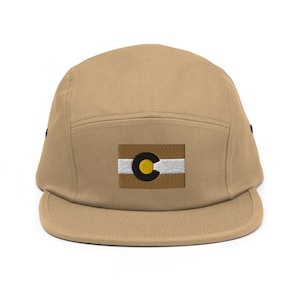 May include: A tan 5-panel cap with a black brim and a white and gold embroidered Colorado flag patch on the front.