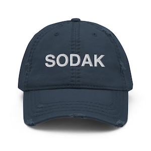 Peut inclure: Une casquette de baseball en denim bleu marine usée avec le mot "SODAK" brodé en blanc sur le devant.
