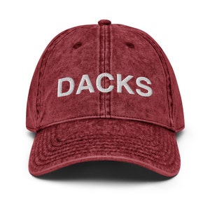 May include: A red vintage washed cotton baseball cap with white embroidered text that reads "DACKS".