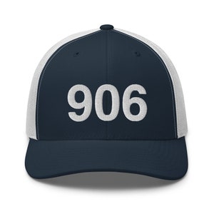 Könnte beinhalten: Marineblaue und weiße Trucker-Cap mit der Zahl "906" in Weiß auf der Vorderseite gestickt. Die Kappe hat einen Mesh-Rücken und einen gebogenen Schirm. Ein klassischer Stil.
