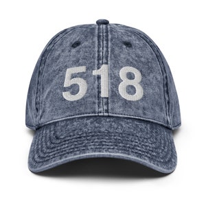 Puede incluir: Una gorra de béisbol de mezclilla azul con el número 518 bordado en blanco en la parte delantera.