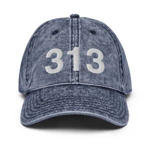 Puede incluir: Una gorra de béisbol de mezclilla azul con el número 313 bordado en blanco en la parte delantera.
