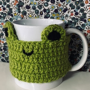 May include: A white ceramic mug with a green crocheted frog-shaped cosy. The cosy has black eyes and a black smile.