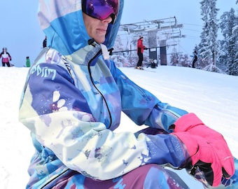 Op de afbeelding: Een persoon draagt een blauw, wit en paars skipak met een roze en zwarte handschoen. De persoon draagt een blauwe capuchontrui met een wit en blauw patroon en een roze skibril.