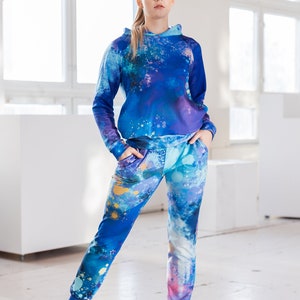 May include: A blue and purple hooded sweatshirt and matching sweatpants with a paint splatter design. The model is wearing pink sneakers.