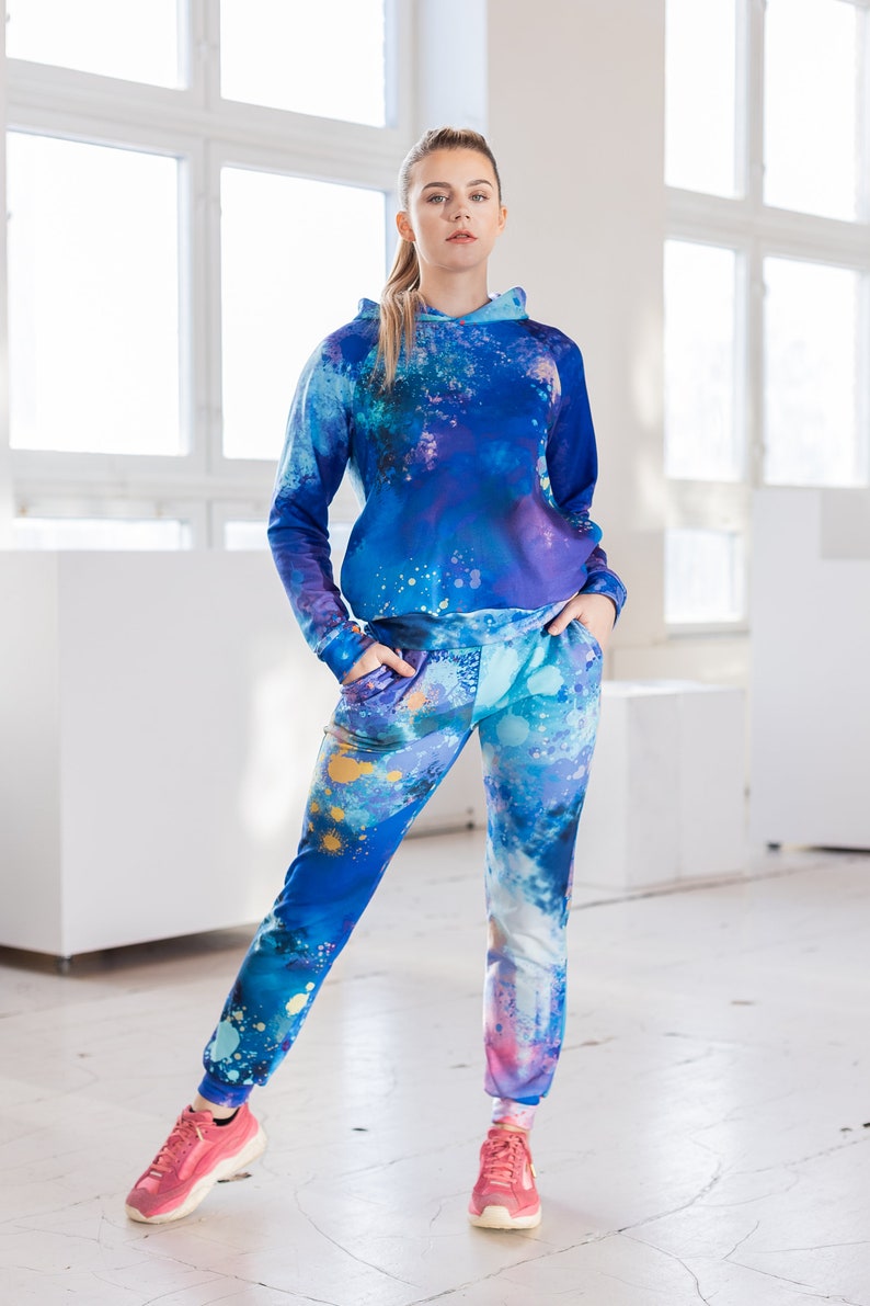 May include: A blue and purple hooded sweatshirt and matching sweatpants with a paint splatter design. The model is wearing pink sneakers.